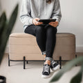 Person in grey hoodie and black jeans sitting on a beige velvet storage ottoman bench with black metal legs, holding a