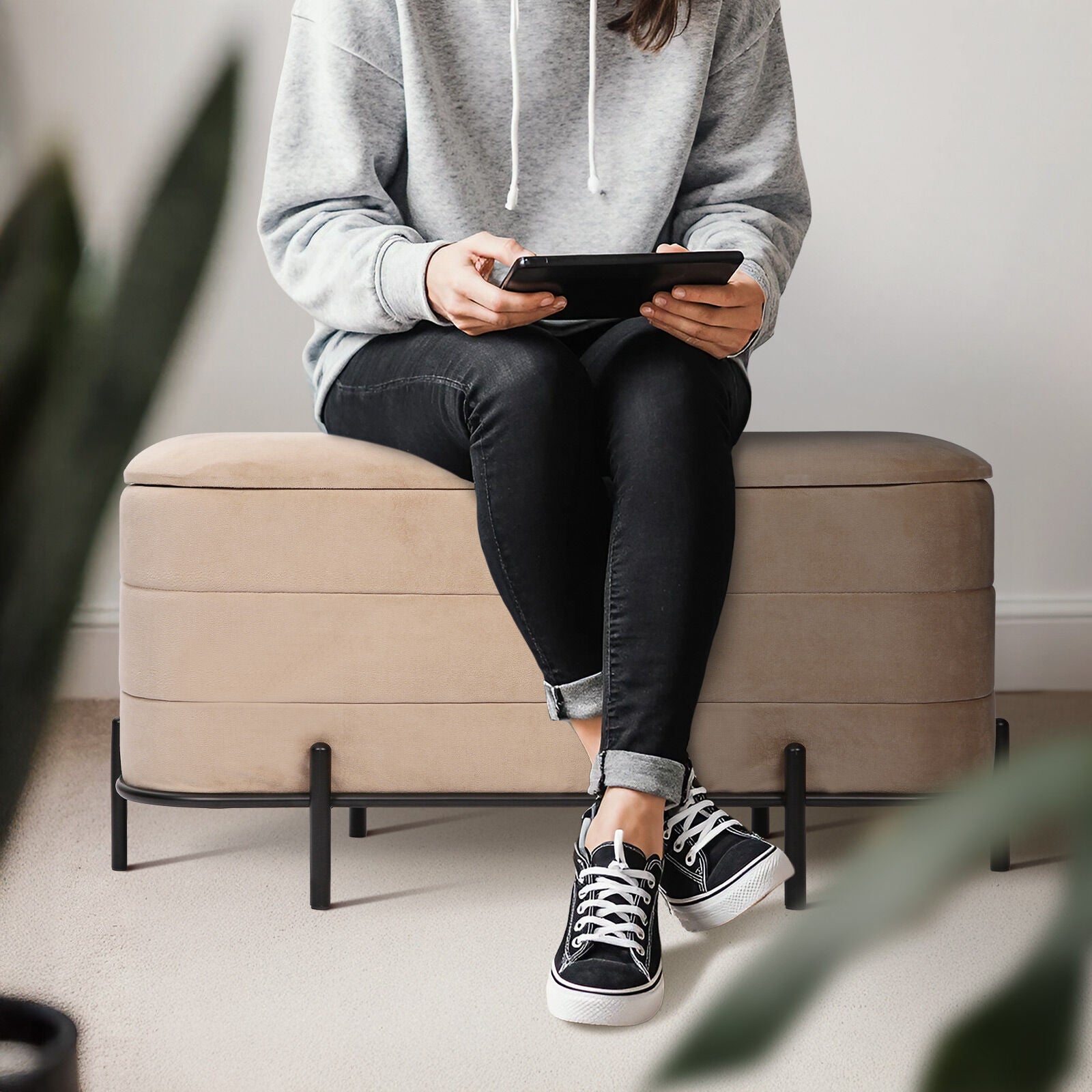 Person in grey hoodie and black jeans sitting on a beige velvet storage ottoman bench with black metal legs, holding a