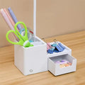Desk lamp organizer base filled with pens, scissors and tape in pen holder, binder clips on shelf, stickers in open drawer