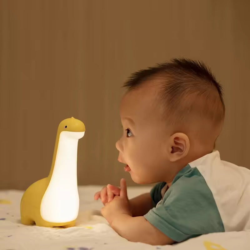 Toddler lying on bed looking at glowing yellow brachiosaurus dinosaur LED night lamp