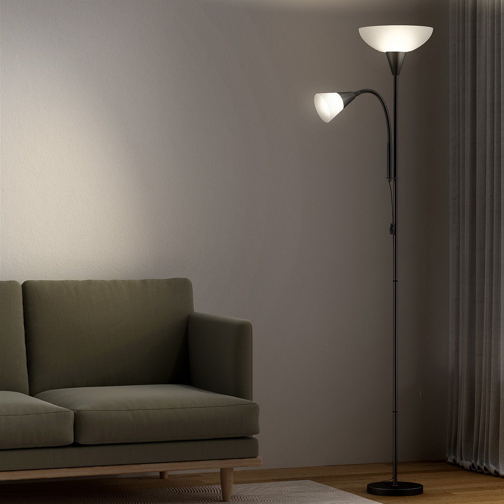 Black dual-shade floor lamp with both shades lit beside dark green sofa in dim living room with curtains