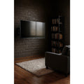 TV wall-mounted on black fixed bracket on dark brick wall lifestyle with grey armchair cream rug dark bookshelf wood floor