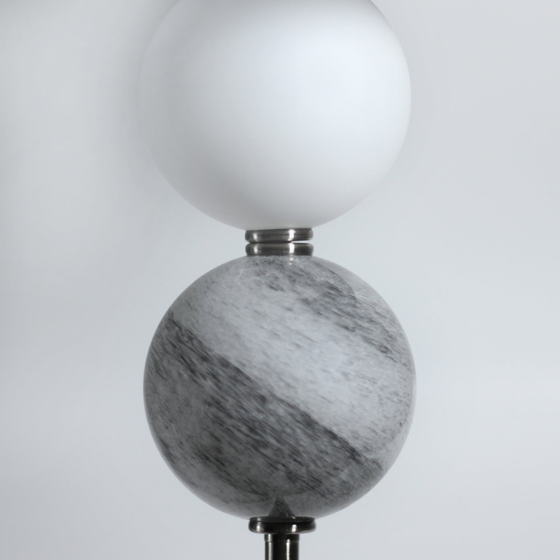 Close-up of white frosted glass ball above larger grey marble-swirled glass ball, chrome collar between, grey background