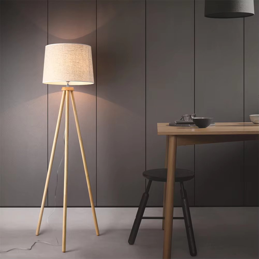 Wood tripod floor lamp with beige linen shade beside grey sofa with cushions, open book on dark wood floor