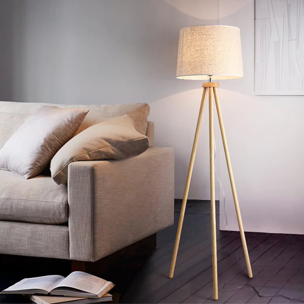Wood tripod floor lamp with beige linen shade beside grey sofa with cushions, open book on dark wood floor