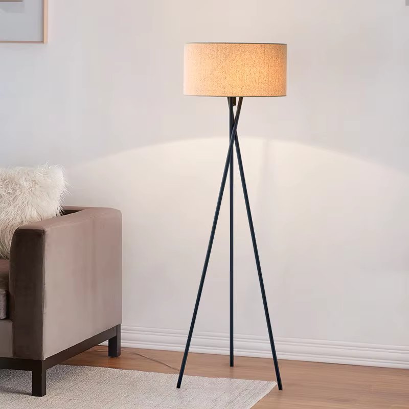 Floor lamp with beige drum shade on dark metal tripod beside brown sofa with fluffy white cushion against white wall