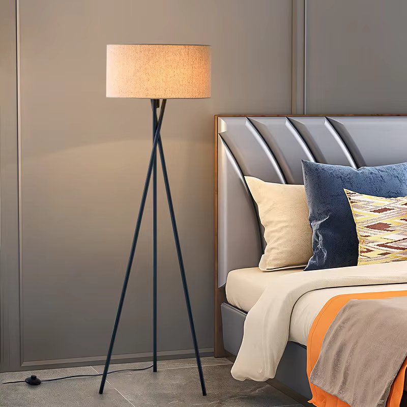 Floor lamp with beige drum shade on dark navy metal tripod base beside upholstered bed with blue and yellow cushions