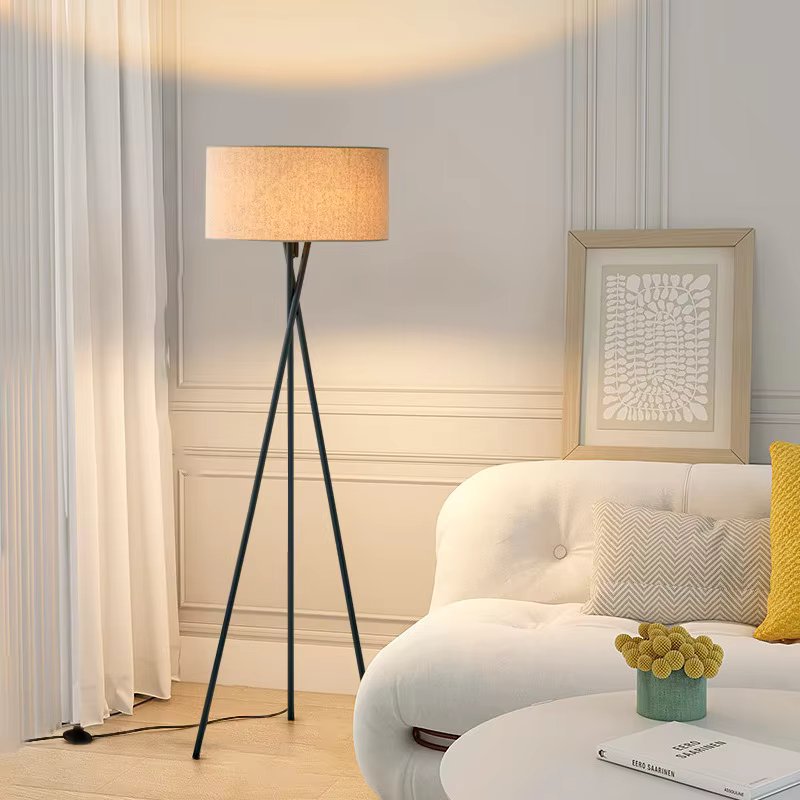 Floor lamp with beige drum shade on dark metal tripod beside white sofa with yellow cushion and artwork in living room