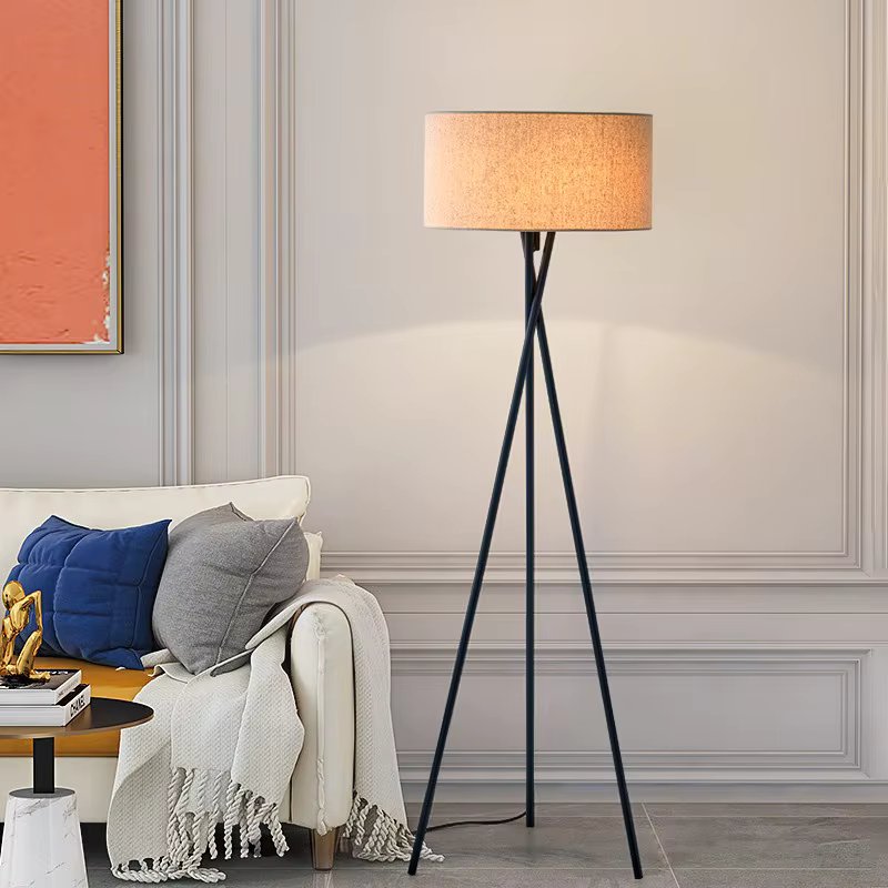 Floor lamp with beige drum shade on dark metal tripod beside white sofa with blue and grey cushions in paneled living room