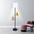 Floor lamp with white drum shade and two round wooden shelves lit in living room, vase on upper shelf, pink armchair beside