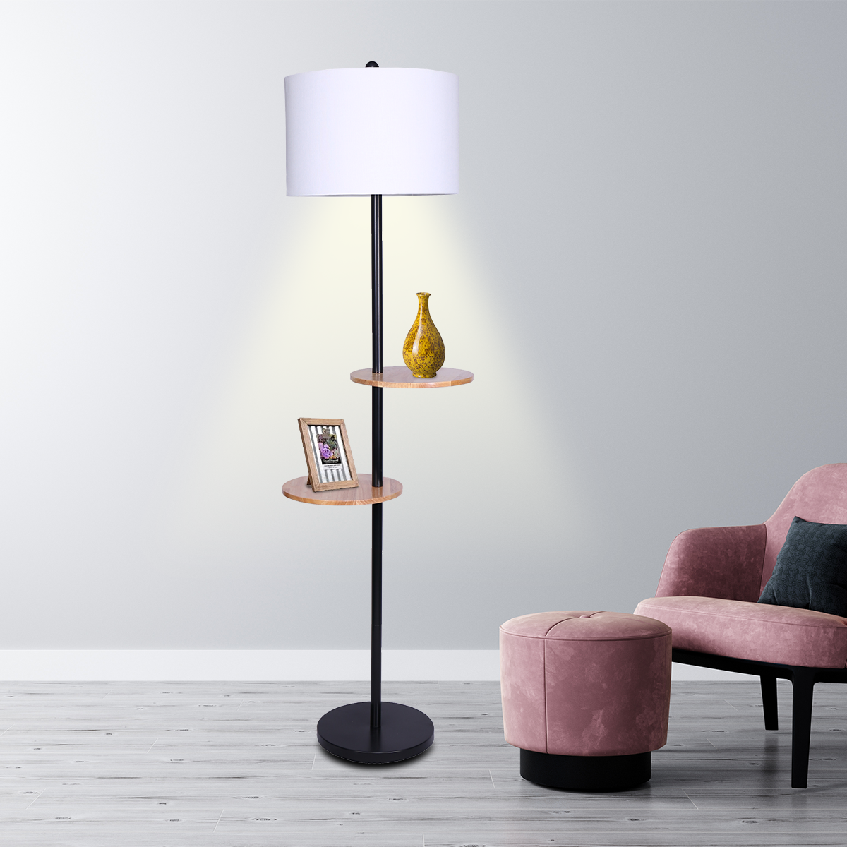 Floor lamp with white drum shade and two round wooden shelves lit in living room, vase on upper shelf, pink armchair beside