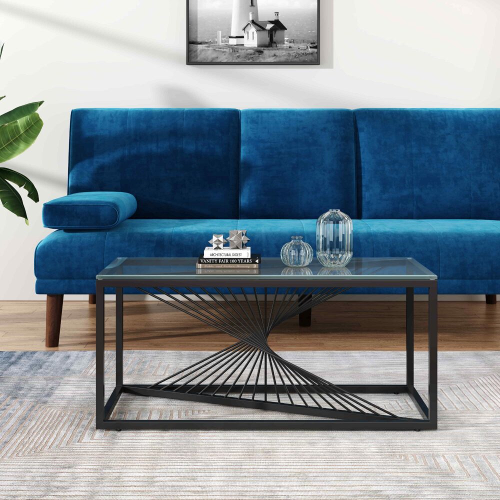 Rectangular glass-top coffee table with black metal starburst base, angled view with blue sofa, potted plant and decorative