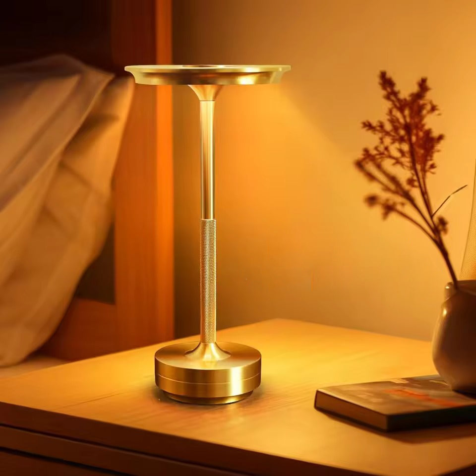 Gold disc-shade lamp glowing warm on bedside table with dried flowers in vase and book beside, warm amber light