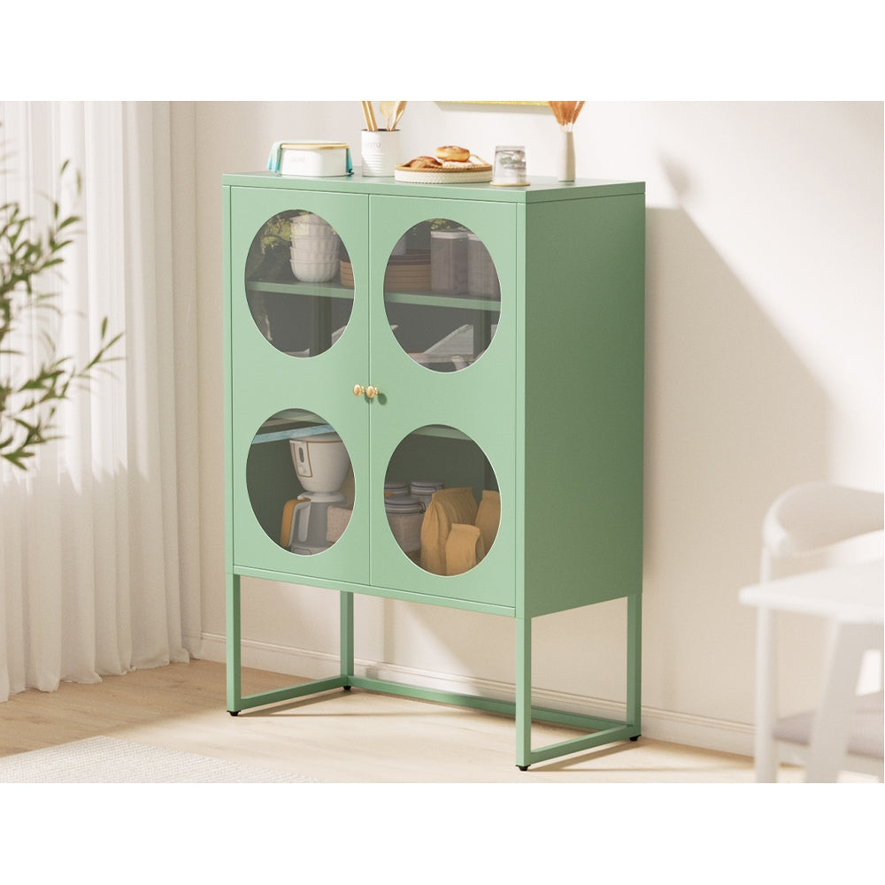 Green metal cabinet with circular glass doors filled with kitchenware, styled in a bright room with white curtains