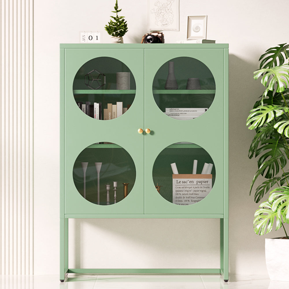 Green metal display cabinet with four circular glass windows and gold knobs, filled with books and decor, in room setting