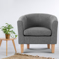 Grey fabric barrel armchair with natural wood legs, monstera plant in copper pot on wooden tripod side table, jute rug