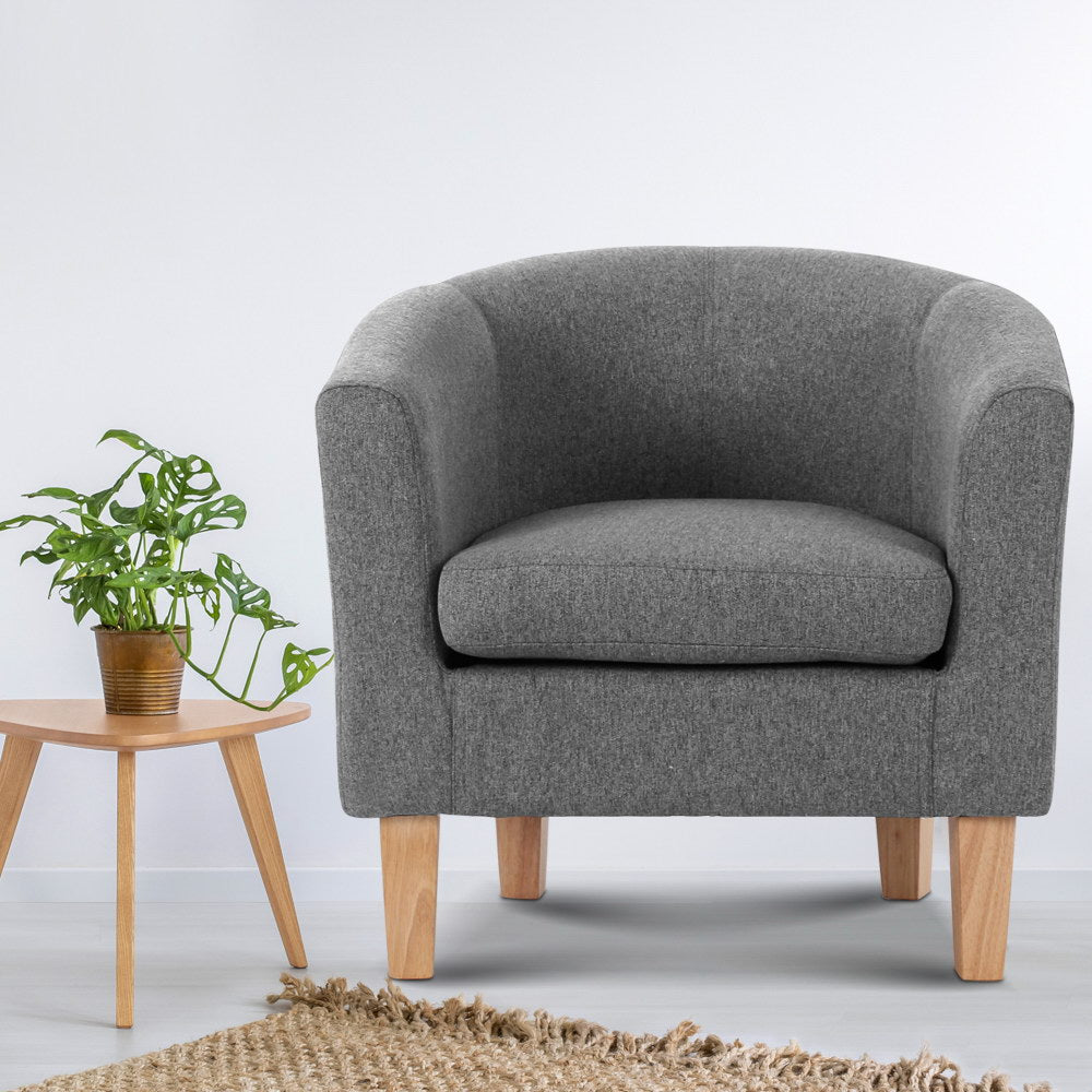 Grey fabric barrel armchair with natural wood legs, monstera plant in copper pot on wooden tripod side table, jute rug