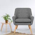 Grey fabric button-tufted armchair with natural wood legs on jute rug, with small round side table and monstera plant beside