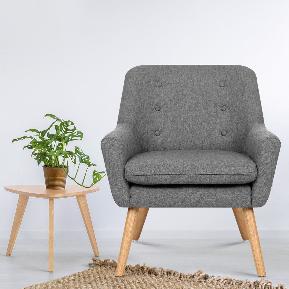 Grey fabric button-tufted armchair with natural wood legs on jute rug, with small round side table and monstera plant beside