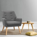 Grey fabric button-tufted armchair with wood legs on striped rug, round wood side table and yellow floor cushion in grey room