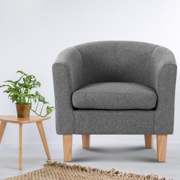 Grey fabric barrel armchair with natural wood legs beside wooden side table with monstera plant, jute rug on white background