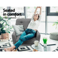 Woman seated sideways in grey fabric button-tufted armchair in bright room, arms raised, 