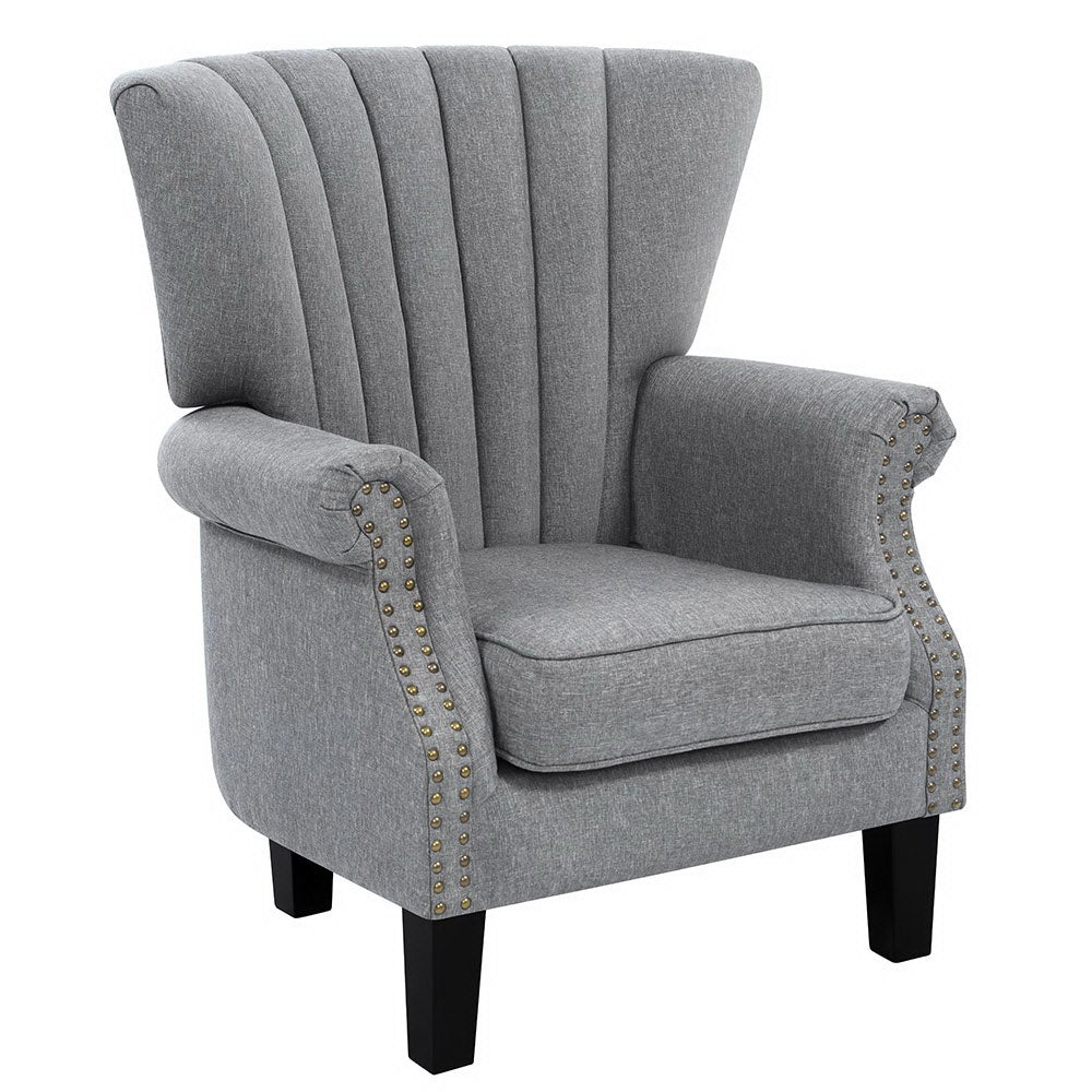 Grey fabric wingback armchair with gold nailhead trim on arms, black legs, side-angle view on white background