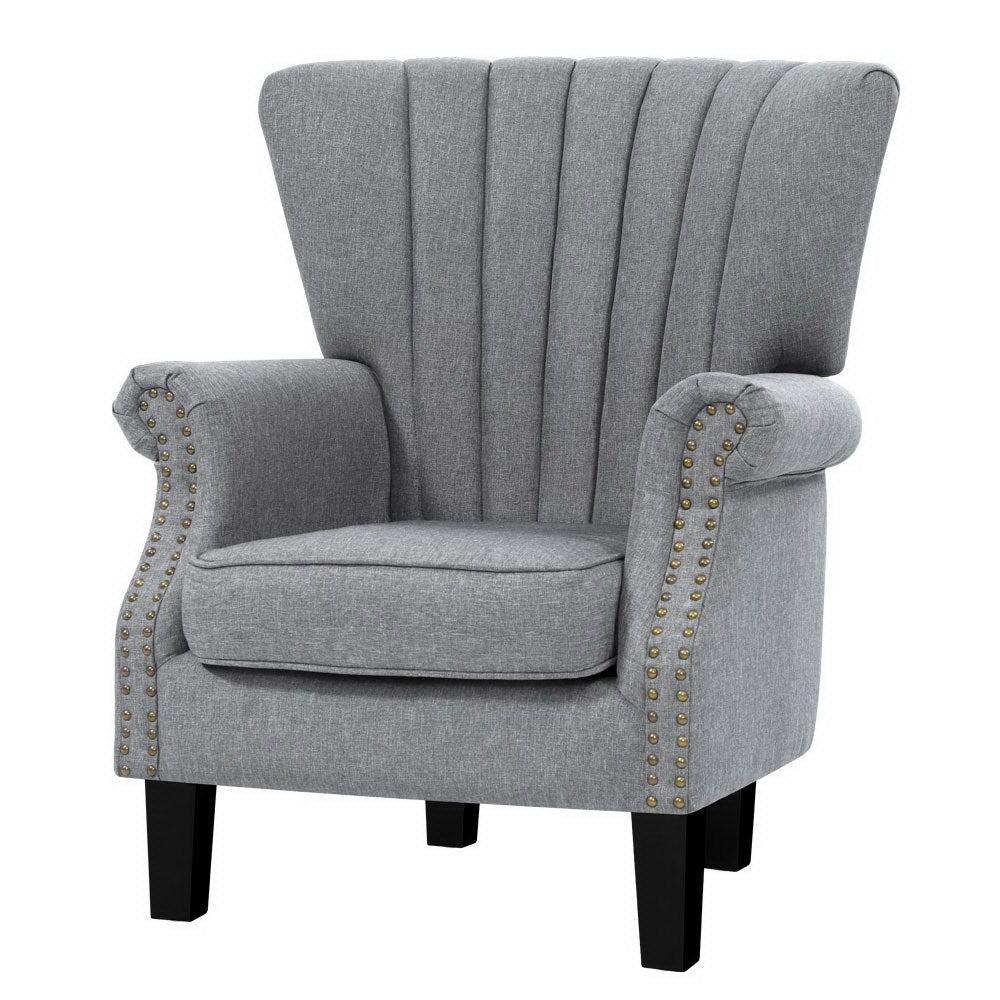 Grey fabric channel-stitched wingback armchair with gold nailhead trim on arms, black legs, angle view on white background