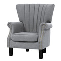 Grey fabric channel-stitched wingback armchair with gold nailhead trim on arms, black legs, angle view on white background