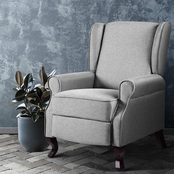 Grey fabric wingback recliner armchair with rolled arms beside rubber plant on brick floor, blue-grey wall
