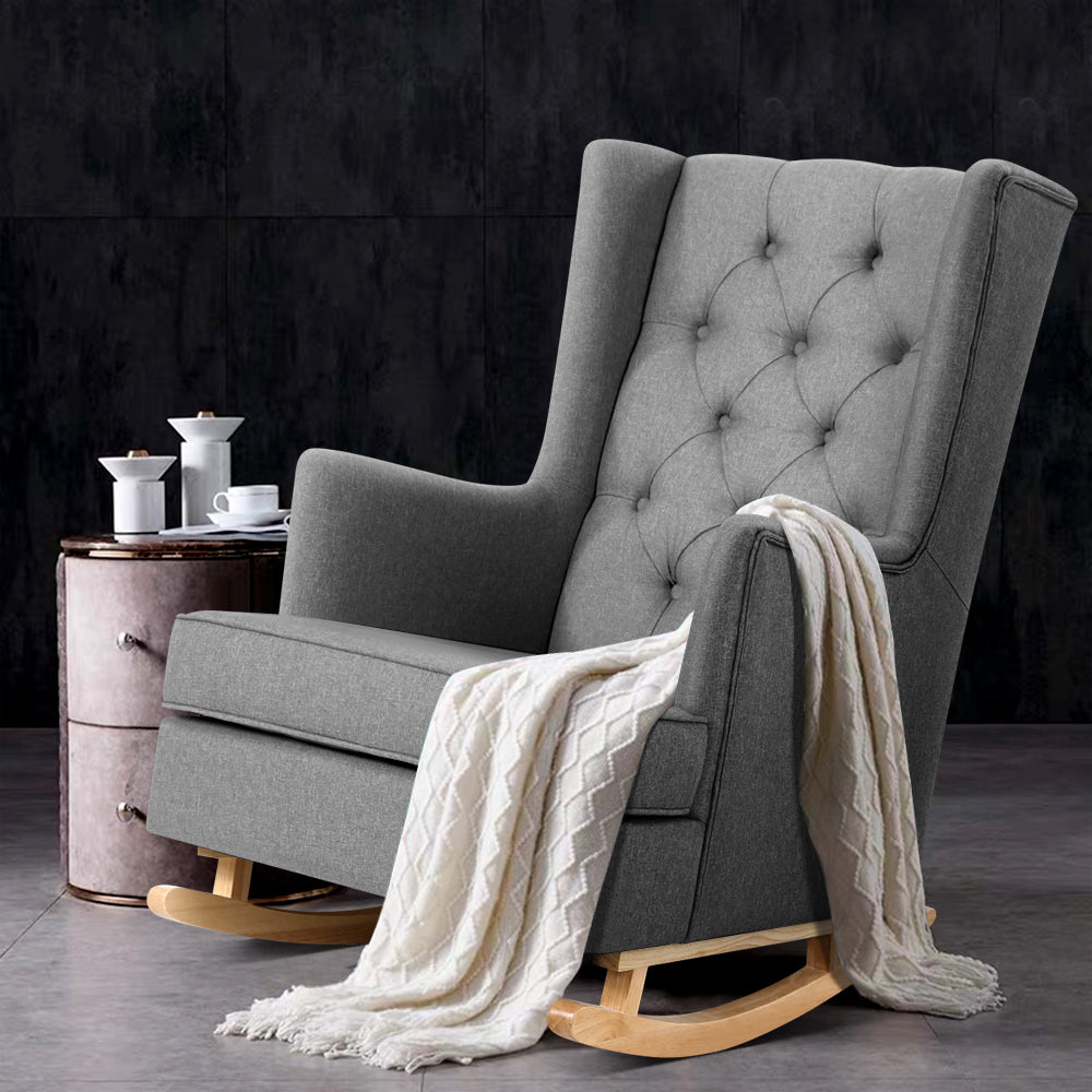 Grey fabric wingback rocking chair with cream knit throw over arm beside vintage side table with tea set, dark wall