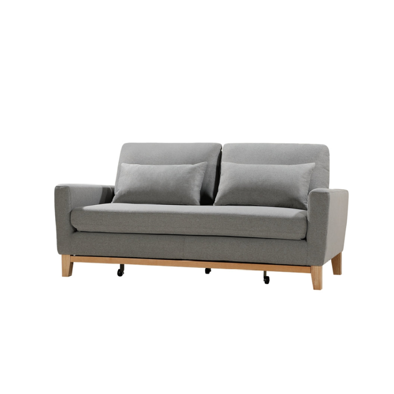 Three-quarter angle view of grey fabric sofa bed with two scatter pillows and wood legs with casters on white background