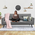 Woman sitting cross-legged on grey fabric sofa bed with laptop and pink throw blanket in styled living room