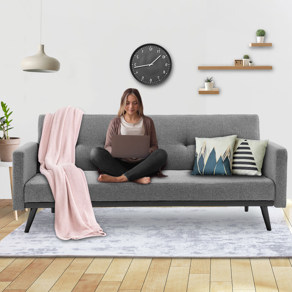 Woman sitting cross-legged on grey fabric sofa bed with laptop and pink throw blanket in styled living room