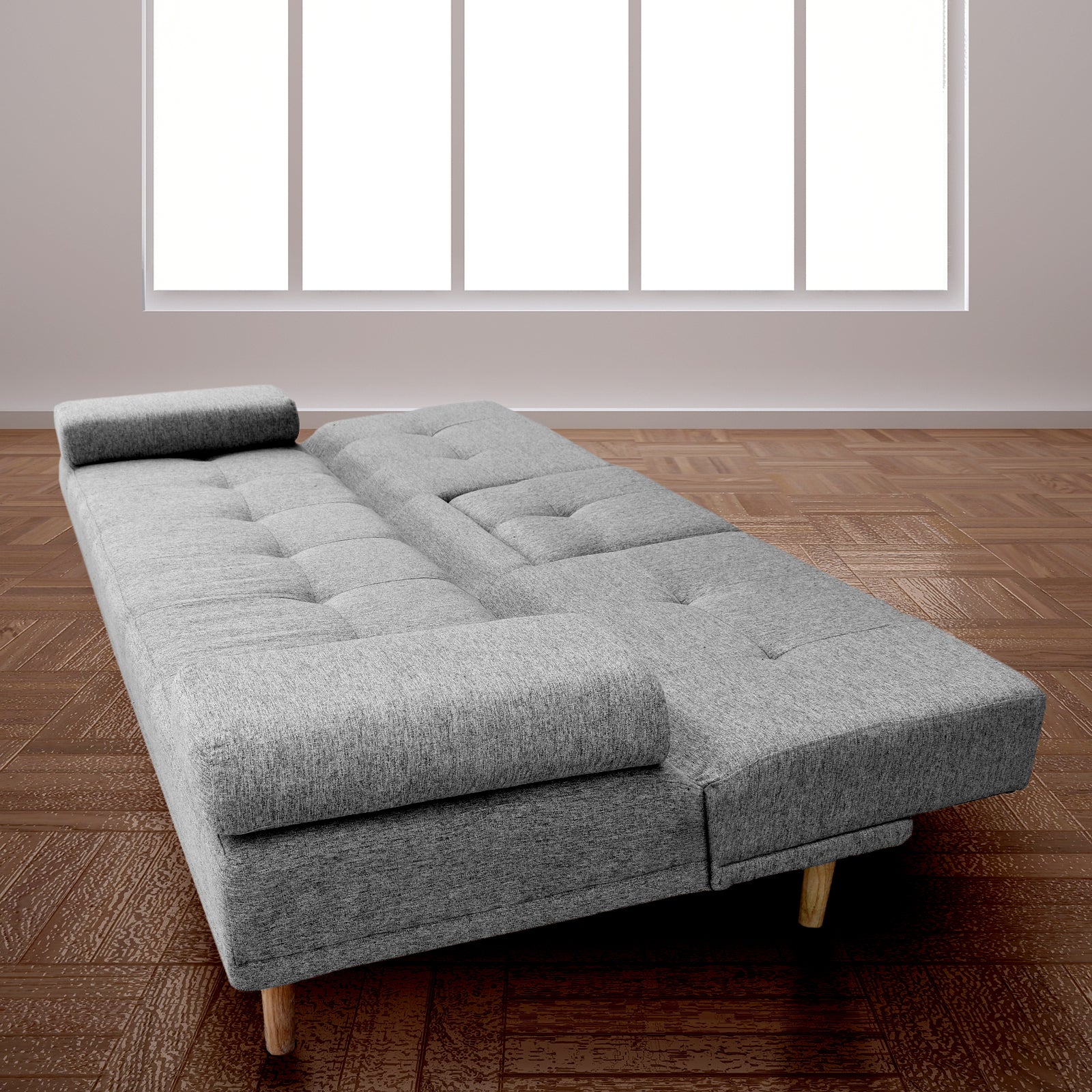 Grey fabric sofa bed fully reclined flat as bed on parquet wood floor by large window with bolster pillow
