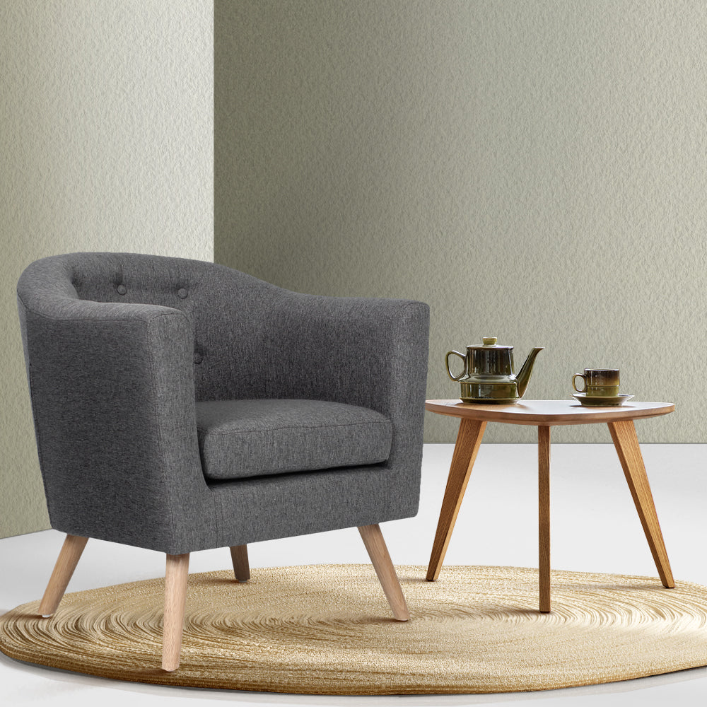 Grey fabric tub armchair with button-tufted back on jute rug beside round wood table with tea set against sage green walls