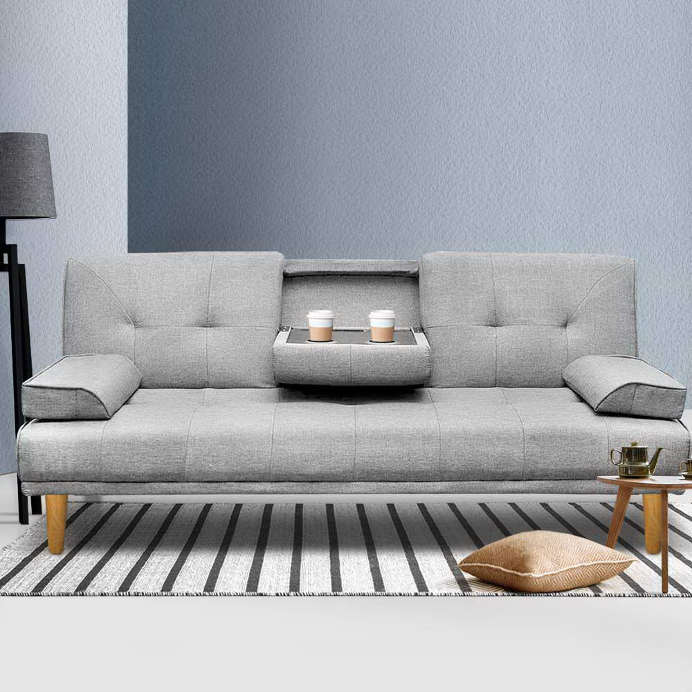 Grey linen sofa bed with cupholder tray holding two coffee cups on striped rug, side table with teapot