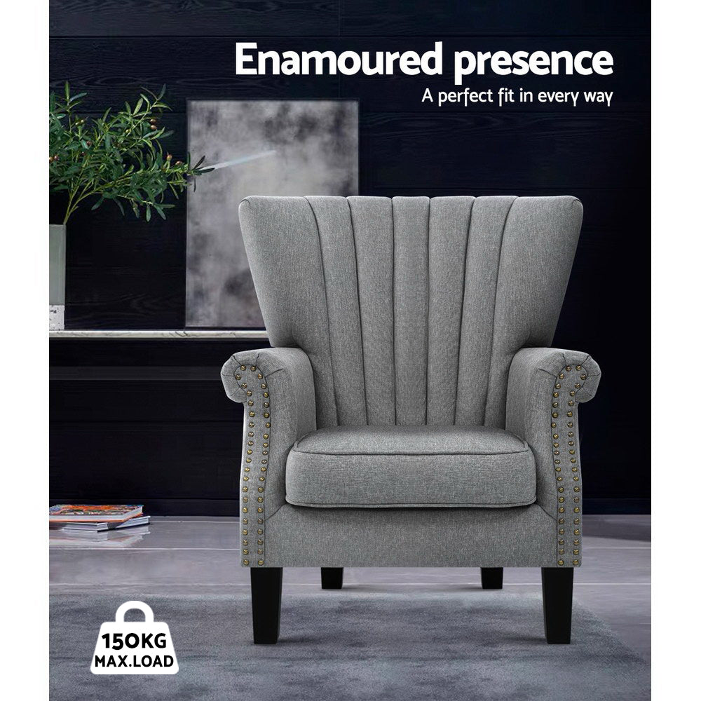 Grey fabric channel-stitched nailhead wingback armchair in dark lifestyle room with plant, 150KG max load badge