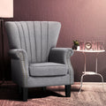 Grey fabric nailhead wingback armchair beside white side table with clock and HOME letters, mauve textured wall background