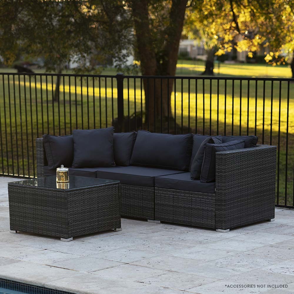 Grey rattan outdoor lounge set with dark cushions and glass coffee table on stone patio beside pool, autumn trees behind