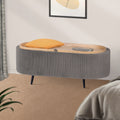 Grey ribbed oval ottoman in bedroom with wood lid partially open, orange pillow and phone resting on lid, beige rug