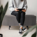 Person seated on grey ribbed oval ottoman holding a tablet, wearing grey hoodie and black jeans