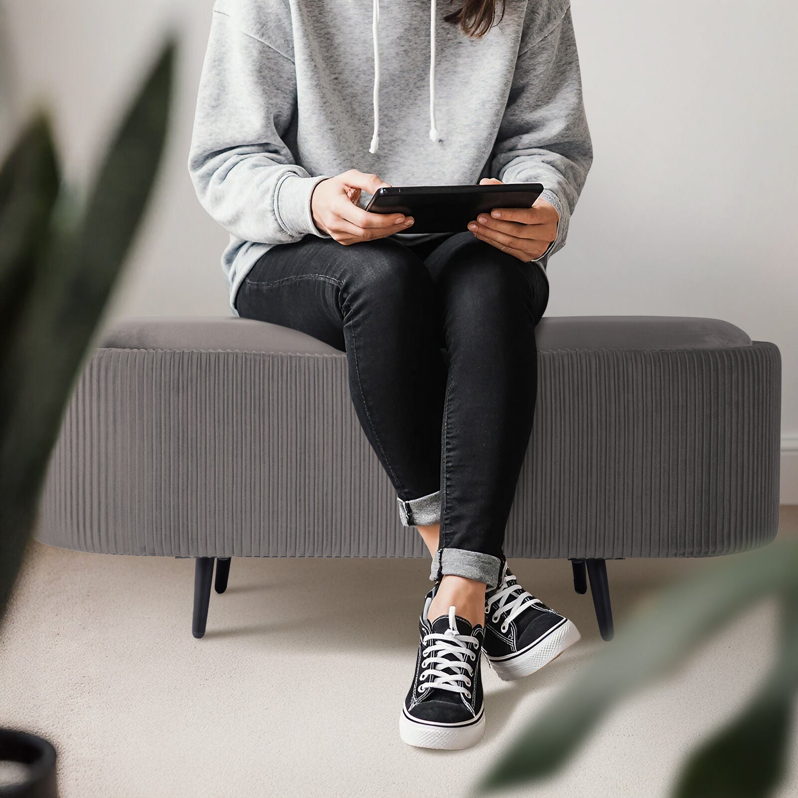 Person seated on grey ribbed oval ottoman holding a tablet, wearing grey hoodie and black jeans