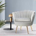 Light grey velvet scallop shell armchair with gold legs beside black side table with gold lamp against light blue wall