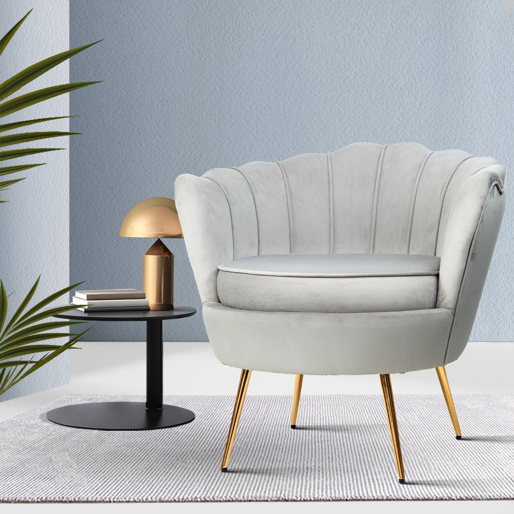 Light grey velvet scallop shell armchair with gold legs beside black side table with gold lamp against light blue wall