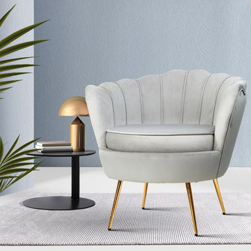 Light grey velvet scallop shell armchair with gold legs beside black side table with gold lamp against light blue wall