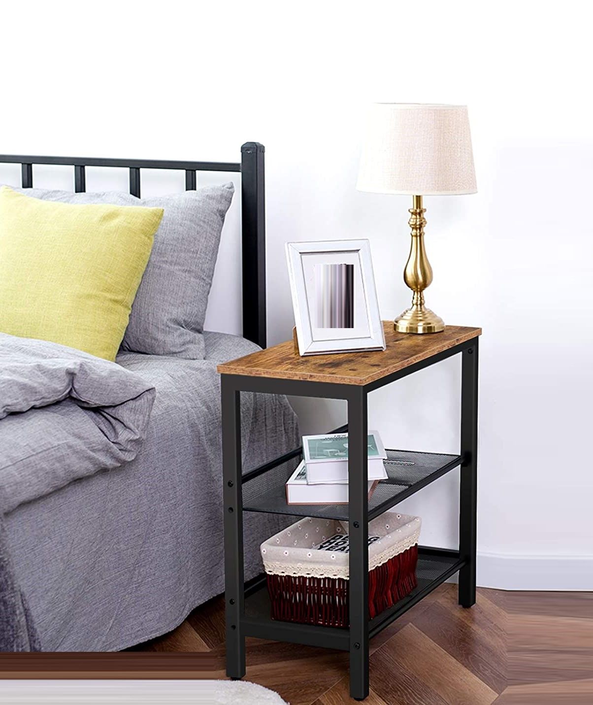 Industrial side table in bedroom beside bed with brass lamp and frame on top, books and basket on mesh shelves