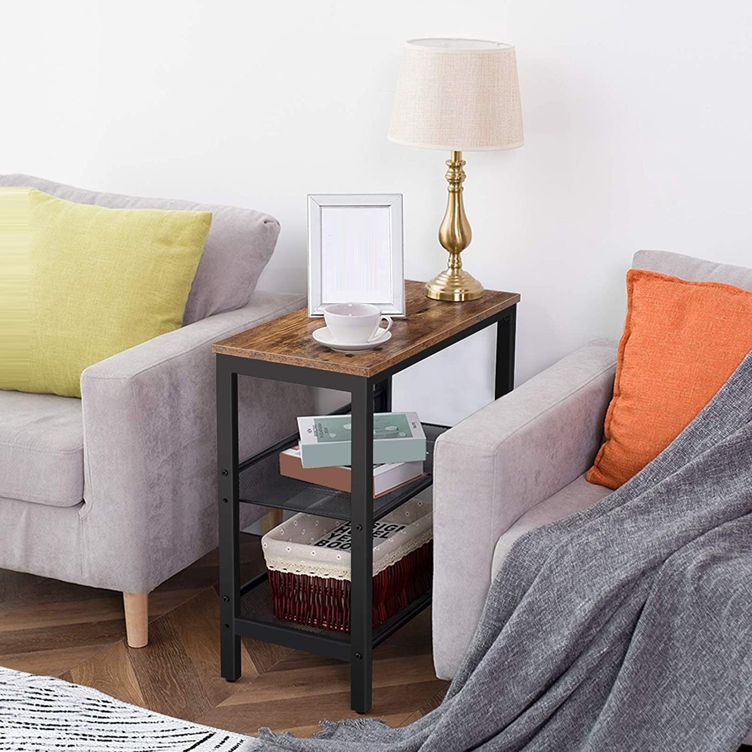 Industrial side table placed between two grey sofas, brass lamp and cup on top, books on lower shelves