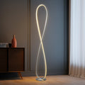 Chrome infinity LED floor lamp glowing warm in dark room, dark grey wall, wood cabinet with terracotta vase and sheer curtain