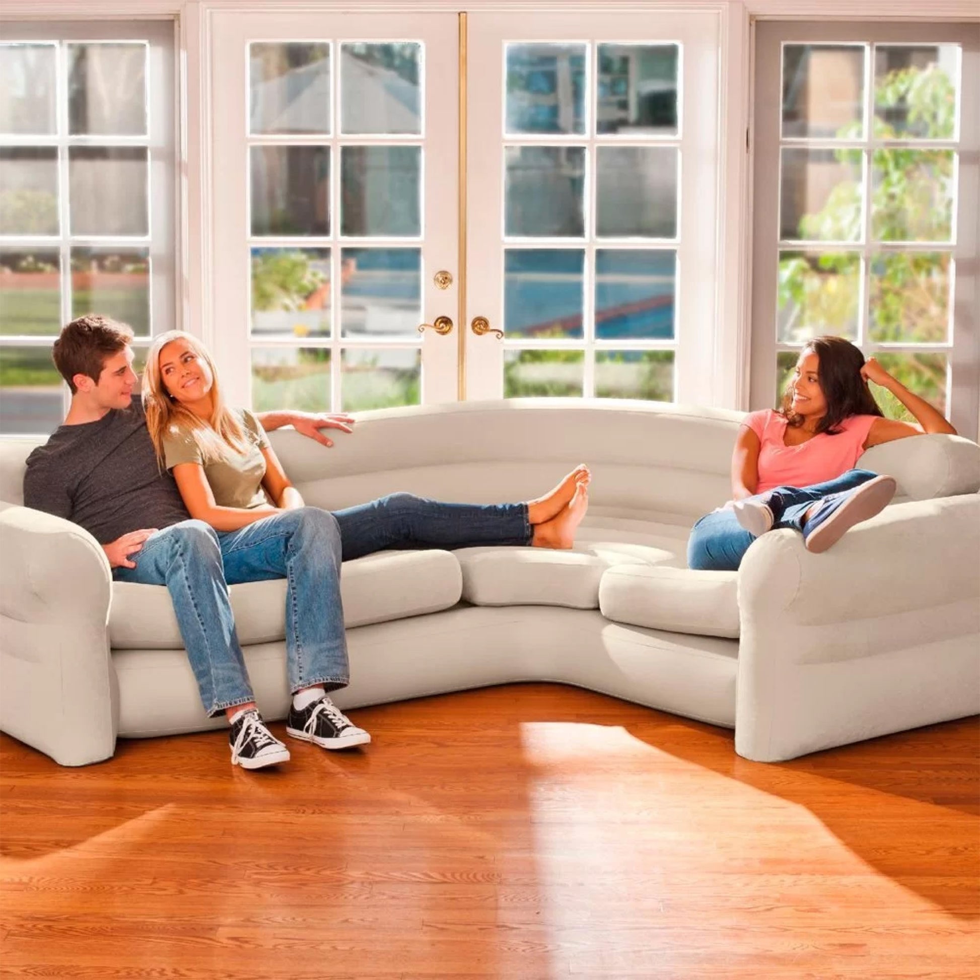 Three people relaxing on beige inflatable corner sofa in bright living room with large patio doors
